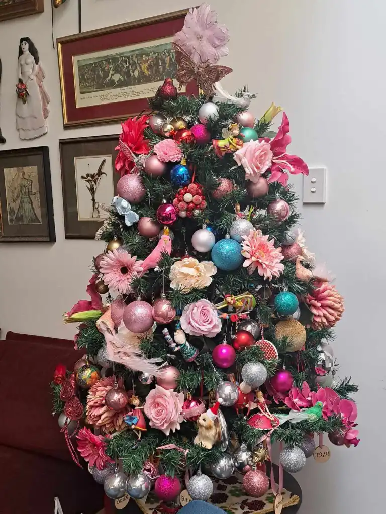 A decorated green Christmas tree (fake not real). Decorated with predominately baubles of various colours, pink flowers and glittery birds